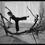 An artist poses in motion that mimics a tree trunk sculpture sitting on the floor. The black and white image shows her reaching above her head and balancing on one foot in a graceful pose.