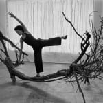 An artist poses in motion that mimics a tree trunk sculpture sitting on the floor. The black and white image shows her reaching above her head and balancing on one foot in a graceful pose.