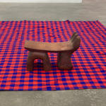 A wooden headrest with three finger-like shapes curving up on one side and three legs on a red and blue patterned cloth