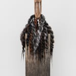 Stylized wood carving of a human figure with locks of hair that resemble a dress around the torso placed on a pedestal