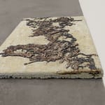 A concrete and bronz sculpture in the size and shape of a sidewalk square sits in a gallery on a concrete floor.