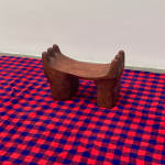 A wooden headrest with thick, stout legs and three finger-like rounded divisions curving up on either side on a red and blue patterned cloth
