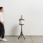 Scale view of figure standing near sculpture with penguin sitting atop of a white pot filled with knick knacks, on wooden pedestal