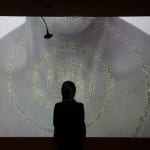 A woman stands in front of a spiral of resin cicadas and a bronze lotus pod projected with a video of the artist's throat