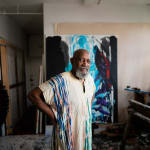 An artist Noah Jemison stands with his hands on his hips surrounded by paintings in a loft setting