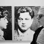 Black and white photograph of Andy Warhol in profile in front of a screen print of a man’s mug shot