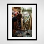 Framed photograph in black wood of an artist, Alex Locadia, stands at a table working on a wood and metal sculpture amidst an industrial loft setting filled with materials and tools