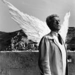 Black and white photograph of Jean Cocteau with feather wings and glass eyes, staring up and to the right in front of a mountain