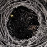Close-up overhead view into a black sculpture reminiscent of a tall birdsnest made of coiled twine revealing a metal jewelry chain and other glistening objects.