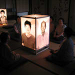 Four people sit around a large illuminated lantern with lifelike photo portraits in a traditional Japanese tatami room