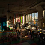 An artist, Steve Silver, standing at a work table amidst paintings and sculptures in an industrial loft setting flooded with late afternoon light
