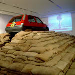 Art installation, a red Renault car on top of a pyramid of sand-filled jute sacks, video projection of atomic explosion