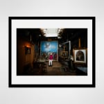 Framed photograph in black wood of an artist, Anne Mason, sitting in front of a large-scale Renaissance-style painting illumnated by a skylight in an otherwise dark loft setting decorated with other paintings