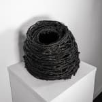 A black sculpture reminiscent of a tall birdsnest made of coiled twine sits on a white pedestal.