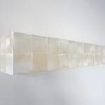 Detail of a row of white iridescent cubes made of acrylic paint on MDF with a pale pink textured strip at its center is mounted on a white wall.