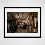 Framed photograph in black wood of an artist, Carmen Cicero, sits at a work desk working on an artwork in an industrial loft setting filled with paintings
