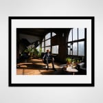 Framed photograph in black wood of an artist, JG Thirlwell, sitting in a chair amidst potted plants next to large arched windows in an industrial loft setting