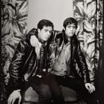 1960s black and white photograph of two young men sitting down with their arms around each other, sitting down and looking to the right wearing black leather jackets