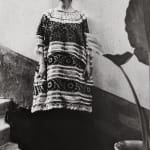 Black and white photograph of Frida Kahlo in traditional Tehuana dress leaning against a bicolored wall next to a plant on the steps outside the Blue House in Mexico City