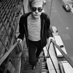 Black and white photograph of Andy Warhol in a Breton striped shirt and sunglasses ascending a fire escape above a city street