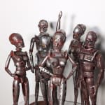 A group of six rust and silver-colored robot figurines, two with male anatomy and four with female anatomy, stand in varying positions on a white pedestal.