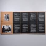Two photographs of an elderly woman, mounted on wood next to eight pages of white text on black paper arranged in a 2x4 grid, hanging on a white wall