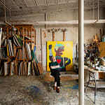 An artist, Carmen Cicero, sits in front of a large figural painting in an industrial loft space filled with stored paintings and materials