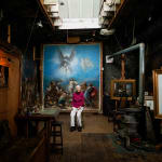 An artist, Anne Mason, sitting in front of a large-scale Renaissance-style painting illumnated by a skylight in an otherwise dark loft setting decorated with other paintings