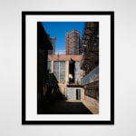 Framed photograph in black wood of an alley view of the exterior of a loft apartment with floor-to-ceiling windows through which an artist, Marsha Pels, peers out