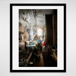 Black framed photograph of an artist, Vernita N'Cognita standing at the end of a hallway in a loft setting filled with plastic containers and other materials