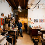 An artist, Joe Haske, leans against a worktable in a loft setting filled with materials and paintings