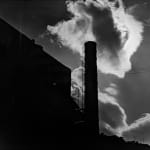 Black and white photograph of the silhouette of a smoke stack against a cloudy sky