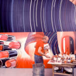 Photograph of James Rosenquist working on a large red and blue painting next to a table with pots of paint