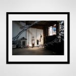Framed photograph in black wood of an artist, Marsha Pels, stands amongst sculptural works in a large industrial loft space