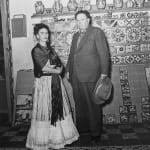 Black and white photograph of Frida Kahlo and Diego Rivera posing in front of a tile-covered wall inside the Blue House