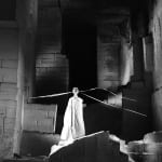 Black and white photograph of “The Oracle” puppet, a bright white figure against buildings ruins