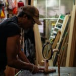 An artist, Alex Locadia, stands at a table working on a wood and metal sculpture amidst an industrial loft setting filled with materials and tools