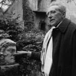 Black and white photograph of Jean Cocteau wearing coat and scarf in his garden and pinching the nose of a classical statue