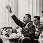 Black & white photograph of Martin Luther King Jr. speaking, right hand raised, people behind him visible