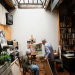 An artist Ken Jacobs standing and artist Flo Jacobs sitting at an easel with an unfinished painting in a loft setting filled with books and illuminated by a skylight