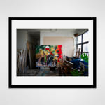 Framed photograph in black wood of a colorful, semi-abstract, figural painting by artist Noah Jemison surrounded by other wrapped paintings in a loft setting
