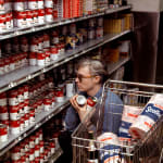 Photograph of Andy Warhol crouching in a grocery store aisle, selecting cans of soup behind a shopping cart