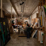 An artist, Carmen Cicero, sits at a desk working on an artwork in an industrial loft setting filled with paintings