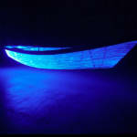 View from the side of a boat made from optic fibers, sitting on a floor in a dark room and emitting a blue light