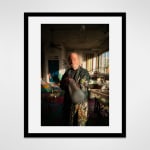 Framed photograph in black wood of an artist, Steve Silver, holding out a cane amidst sculptures and paintings in an industrial loft setting flooded with late afternoon light