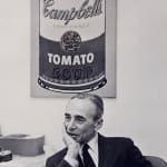 Black and white photograph of Leo Castelli sitting in an office in front of a Warhol soup can painting