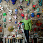 An artist, Jeff Way, poses on a ladder in front of a wall of colorful masks in a loft setting