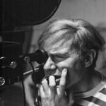 Black and white photograph of Andy Warhol looking into a film camera and biting his nails