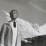 Black and white photograph of Jean Cocteau with glass eyes outside looking up and to the left with the sphinx puppet spreading its wings and looking to the right behind him
