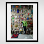 Black framed photograph of an artist, Jeff Way, poses on a ladder in front of a wall of colorful masks in a loft setting